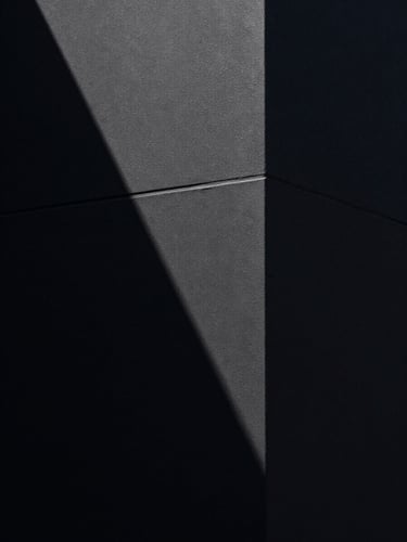 Extreme close-up of a matte black architectural wall corner under dramatic side-lighting, deep chiaroscuro shadows carving a precise vertical line, no fill light, the texture of the surface absorbing all ambient glow — empty, monumental, still