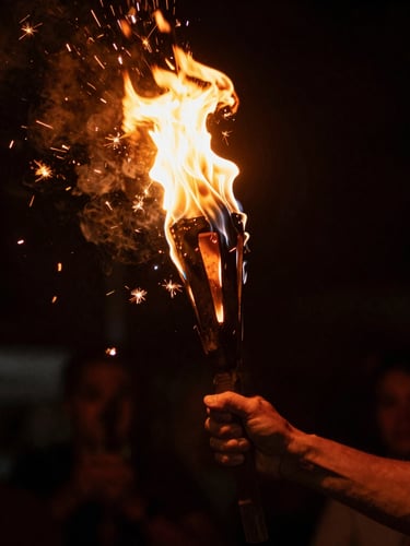 Low-key, high-contrast close-up of hands holding a burning torch during a traditional solstice festival, dramatic orange sparks flying against a deep black background, warm solar flares.