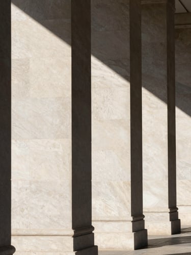 Direct sunlight casting sharp geometric shadows across monolithic travertine columns, architectural elevation, high contrast, clean minimalist lines