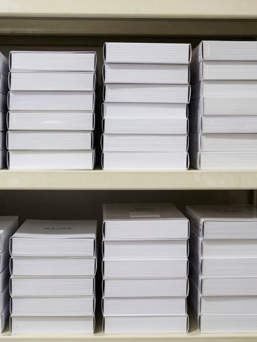 Straight-on flatlay of neatly stacked boxes of commercial stationery and order pads on clean warehouse shelving, bright high-key commercial lighting.