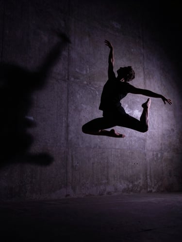 Dramatic low-angle cinematic editorial photography of a dancer in mid-air silhouette against a dark, high-contrast backlit concrete wall, deep shadows, 35mm anamorphic lens.