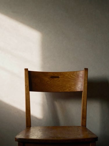 A quiet interior space with a single wooden chair, soft directional north-facing window light casting long shadows on a warm gray wall.