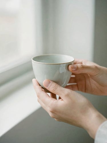 A close-up of a person's hands gently holding a warm, hand-thrown ceramic cup, soft morning daylight diffusing through a nearby window, minimalist composition, 35mm film look.