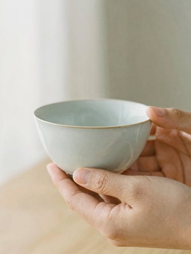 Close-up of hands gently holding a warm, hand-thrown ceramic tea bowl, soft morning daylight, warm neutral tones, minimalist composition