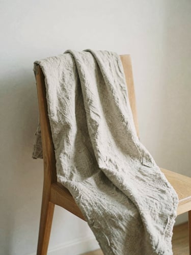 Soft diffused morning daylight illuminating a textured organic linen fabric draped over a light oak chair, warm neutral tones, minimalist composition, 35mm film look