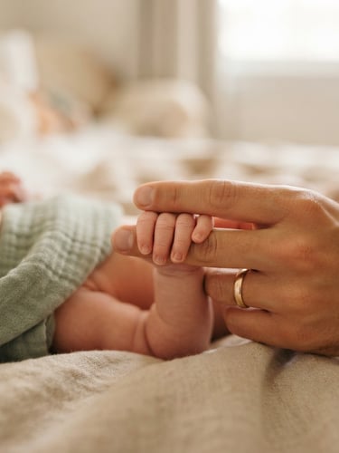 Close-up of a parent's hand gently holding a newborn's tiny fingers, soft morning daylight, warm beige background, shallow depth of field, 35mm