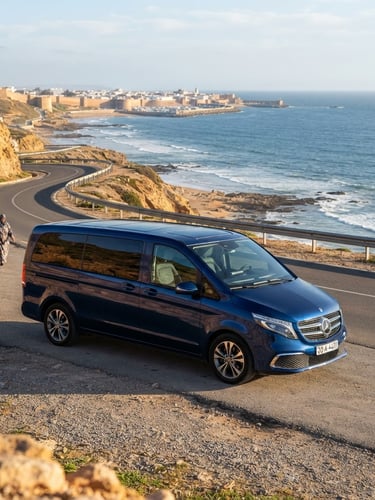A spotless modern Mercedes-Benz V-Class van parked along the scenic coastal highway of Essaouira, bright natural daylight reflecting off the polished deep blue exterior, Atlantic ocean in the background.