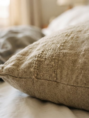 Macro shot of thick oatmeal linen pillow cover on a relaxed unmade bed, soft morning window light, shallow depth of field