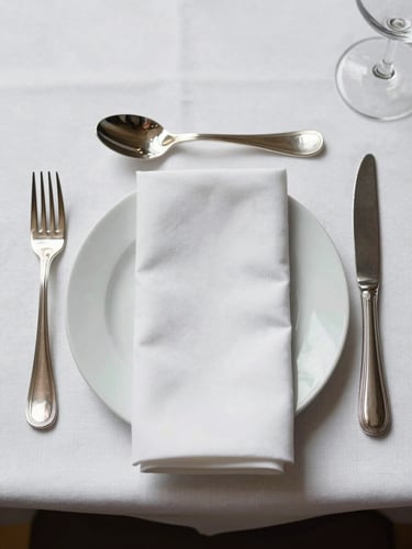 Overhead close study of a formal place setting on a white linen tablecloth — bone china plate, polished silver cutlery aligned precisely, a single folded napkin, shadowless studio light, no food, no people, geometry of the arrangement is the subject