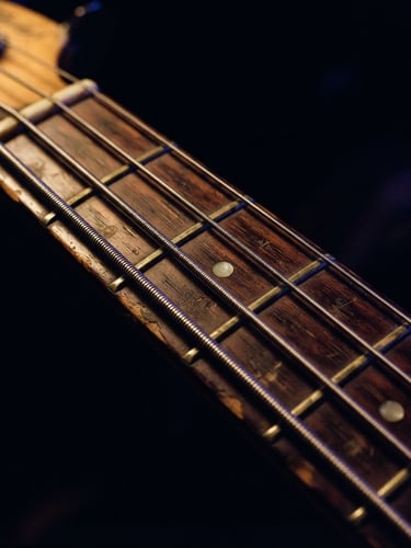 Extreme close-up of a vintage bass guitar neck, warm tungsten glow on worn wood and metal strings, deep obsidian background, 35mm film grain.