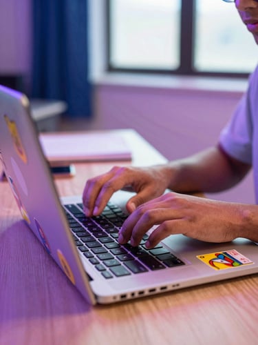 Vibrant close-up of a student typing on a laptop covered in colorful stickers under energetic neon desk lighting, shallow depth of field, 35mm