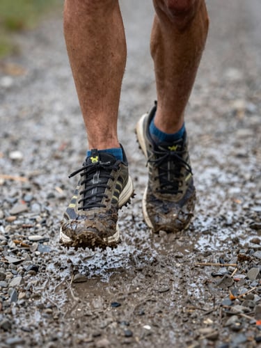 Dynamic close-up of muddy trail running shoes mid-stride on wet gravel, high-contrast natural daylight, shallow depth of field, 35mm