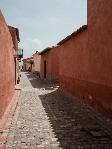 Wide environmental shot of a narrow Mediterranean village lane at midday, aged terracotta walls casting a long shadow across worn cobblestones, empty, still, soft natural light, framed from street level with sky visible above