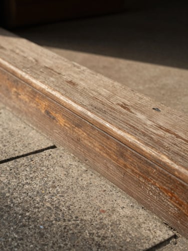 Close-up of a weathered wooden threshold and stone doorstep bathed in soft noon light, worn surface texture visible, no people, warm diffused shadows, eye-level framing with generous negative space to the left