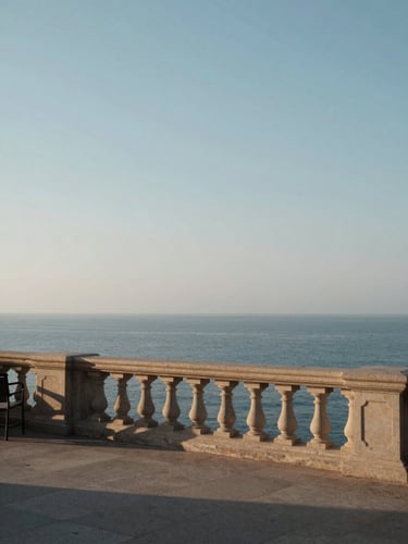 Wide stone terrace railing overlooking a still blue-grey sea, empty chair to the far left edge, morning haze softening the horizon, eye-level framing with generous sky above