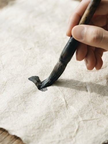 Extreme close-up flat lay of hands holding a wet bamboo calligraphy brush over raw, textured Xuan paper, soft natural side-light casting gentle shadows.