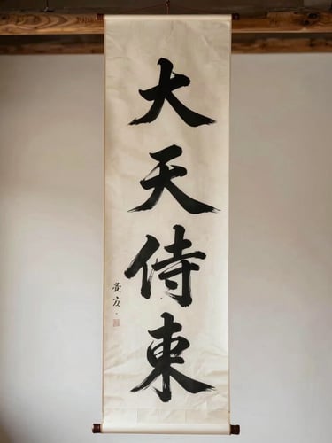 A tall vertical paper scroll with bold black calligraphy hanging in a serene, sunlit room with wooden beams.