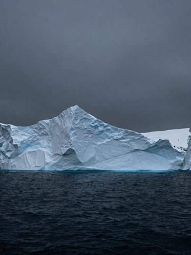 A dramatic vertical portrait of a monolithic iceberg in a dark charcoal arctic ocean, cold overcast daylight, high contrast, cinematic wide-angle.