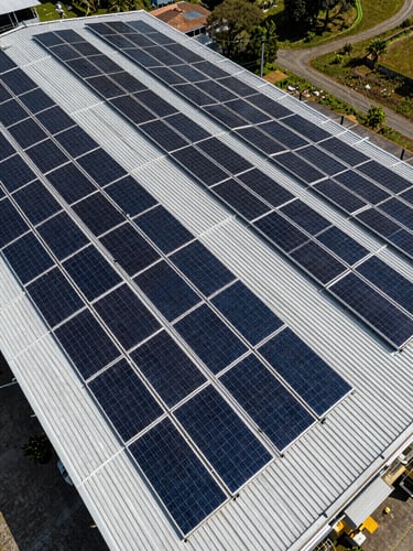 Aerial drone shot of a massive industrial metal roof in Colombia covered with neat rows of monocrystalline solar panels under bright, direct sunlight, sharp geometric shadows.