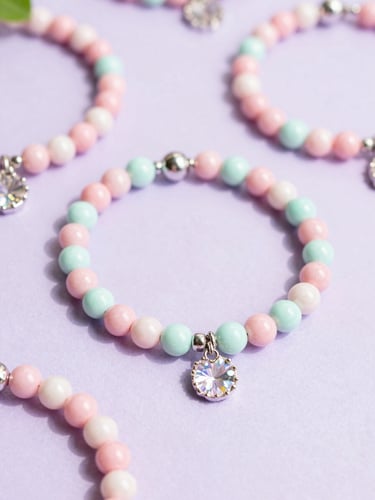 Bright pastel flatlay of iridescent pink and mint beaded bracelets on a soft lilac background, natural morning daylight, sparkling charms.