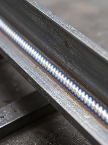Extreme close-up of a freshly completed MIG weld bead on a structural steel I-beam, shot from low angle with hard industrial daylight raking across the surface from the left, revealing the precise ripple pattern of the joint and the surrounding heat-affected zone — no people, no background detail, just the weld and the steel