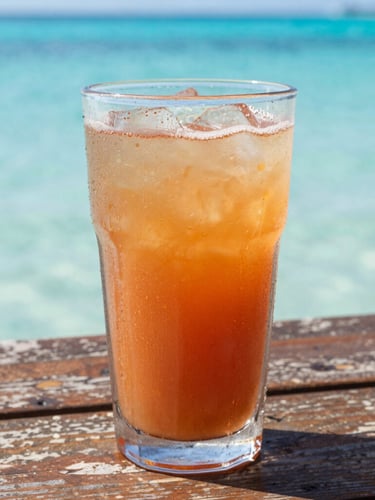 A close-up details shot of a cold tropical drink with condensation on the glass, sitting on a weathered wooden table with bright turquoise ocean water blurred in the background, warm golden sunlight