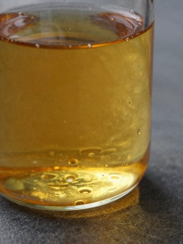 Extreme close-up of amber castor oil filling a clear glass vessel, north-facing daylight streaming through the liquid, visible light refraction casting warm ochre tones on a dark stone surface, tight frame with the glass edge bleeding right