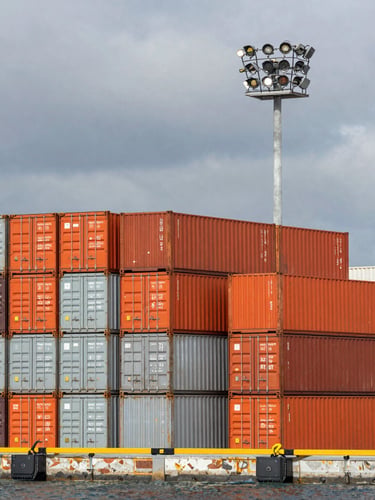 High-contrast wide shot of a modern deep-water port terminal with stacked orange and gray shipping containers, overcast sky, sharp industrial lighting.