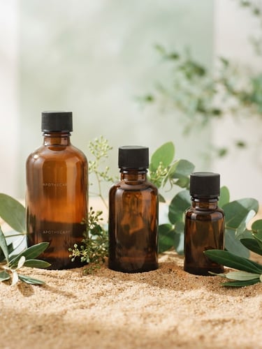 Minimalist studio shot of amber glass apothecary bottles and green leaves on a warm sand surface, soft morning light, clean shadows