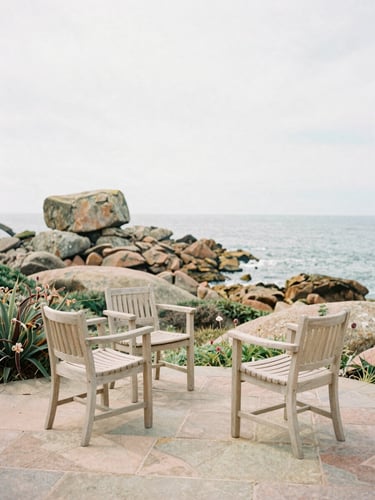 A quiet stone terrace with light wooden chairs, overlooking a wild coastal garden with pink granite rocks and sea views, soft overcast Breton daylight, warm editorial photography, medium format film