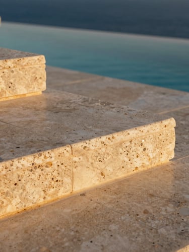 Close-up of raw travertine stone terrace tiles under low-angle golden hour sun, water pool edge blurring into the deep blue sea horizon.
