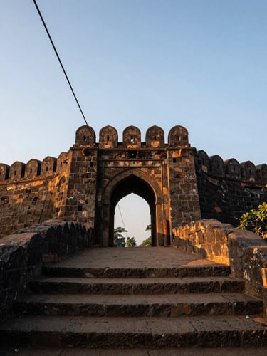 Wide environmental shot looking up at Raigad Fort stone gateway at dawn, ropeway cable visible in upper left corner, rough laterite steps in foreground, golden-hour rim light catching the top of the battlements, deep shadow in the archway, sparse vegetation on the hillside, no tourists visible