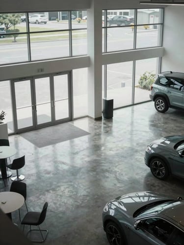 A high-angle architectural shot of a modern, clean dealership showroom, polished concrete floors reflecting overcast daylight through massive glass windows, minimalist aesthetic.