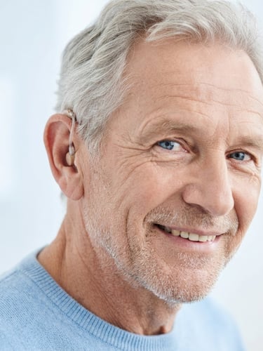 Discreet close-up portrait of an active senior smiling, wearing a near-invisible behind-the-ear hearing aid, bright high-key clinical studio lighting, soft blue tones, 85mm