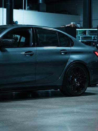 Low-angle profile of a dark grey BMW M-series parked in a clean, modern workshop bay, cool blue ambient light highlighting its metallic paint, with a blurred diagnostic screen displaying code in the background. High-contrast studio lighting.