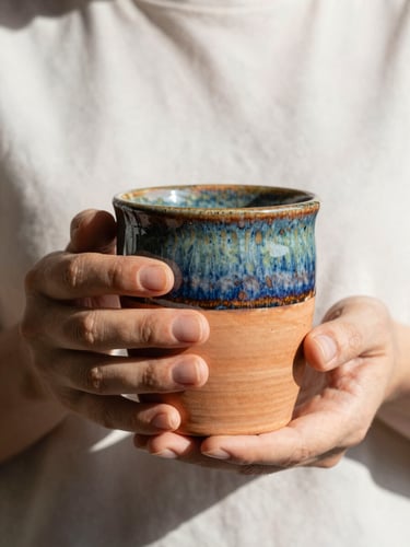 A portrait shot of a person holding a warm terracotta stoneware mug with both hands, soft morning daylight, rich colorful glazes, tactile stoneware textures, 35mm