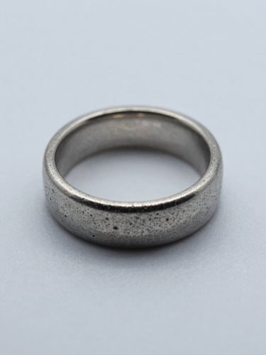 Straight-on close-up of a finished cast metal ring on a flat matte surface, cool even studio daylight, shadowless white context, precise metal grain and edge detail visible, no hands or props