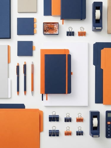 Clean flatlay of industrial office stationery, including neatly stacked files, folders, notebooks, and printer paper, arranged with sharp lines under bright studio lighting. Navy blue and orange accents visible.