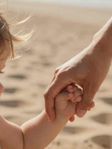 A close-up macro shot of a mother holding her toddler's hand, warm low-angle sunlight highlighting wind-blown hair, soft sand textures in the background.