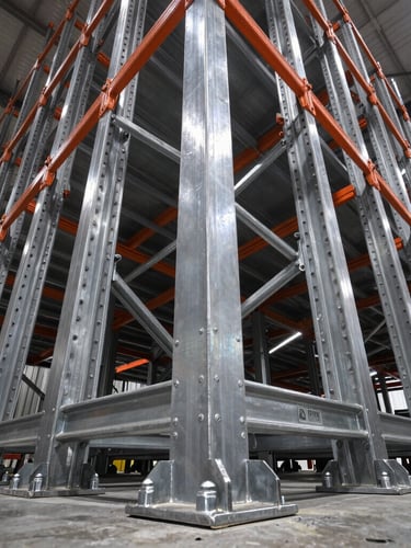 Low-angle shot looking up at massive industrial racking uprights, sharp focus on heavy-duty steel welds and bolted baseplates, high-contrast warehouse lighting, cool steel tones.