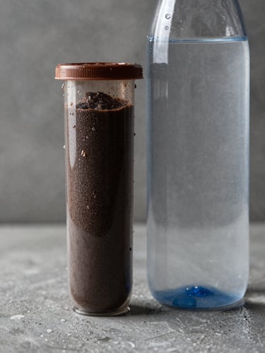Macro shot of a dark-textured protein tub next to a high-pH alkaline water bottle, dramatic side lighting defining sharp edges and water droplets, industrial concrete background