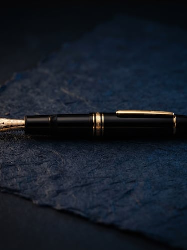 Macro studio profile of a polished luxury fountain pen resting on heavy textured paper, dramatic low-key gold and navy lighting, shallow depth of field