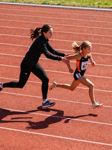 A side-profile shot of a female coach encouraging a young runner on a track, high contrast, bright morning sun, dynamic motion.