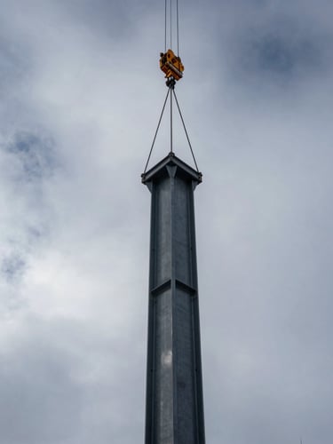 Low-angle shot of a massive structural steel column being hoisted by a crane against a dramatic, overcast Wyoming winter sky.