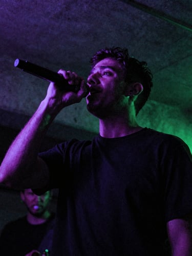 Low-angle gritty portrait of SergioMdrz performing in a dark concrete basement, glowing violet and neon green gel lights, motion blur, 35mm