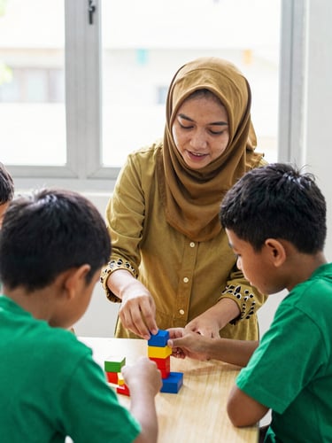 A warm, candid photo of an Indonesian teacher guiding two elementary students as they work together on a simple science project with colorful blocks near a window with soft light.