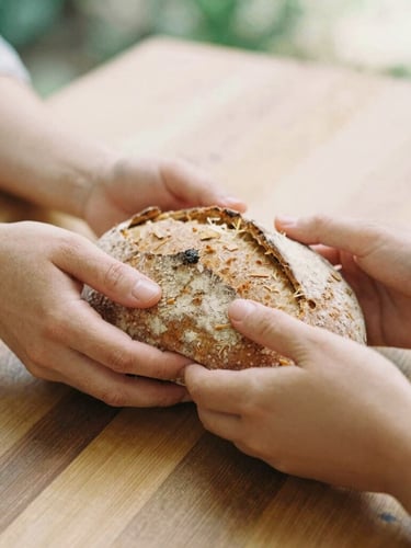 Warm editorial photography, soft natural daylight, close-up of hands sharing rustic bread over a wooden table, earthy organic tones, 35mm film look