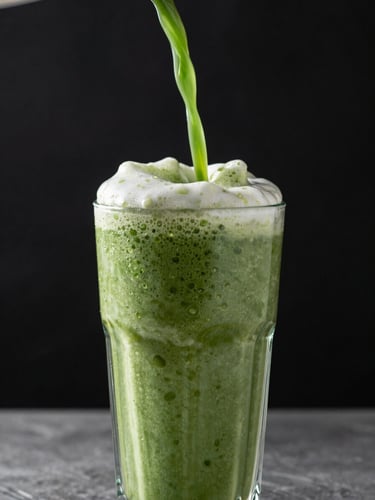 Close-up vertical frame of a green Herbalife batido being poured into a tall glass, vibrant emerald foam cresting at the rim, studio strobe catching droplets mid-air against deep black background, blurred stainless counter below