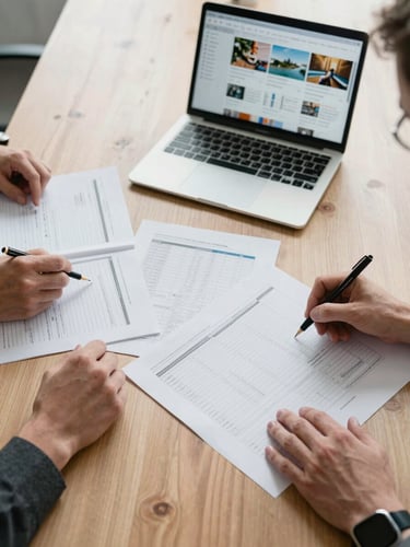 Wide overhead flat-lay view of a strategy session in progress — a campaign brief spread across a conference table, a laptop showing a media plan dashboard, and two sets of hands annotating a printed channel grid under clean office daylight. No faces visible, focus tight on the working documents and screen.