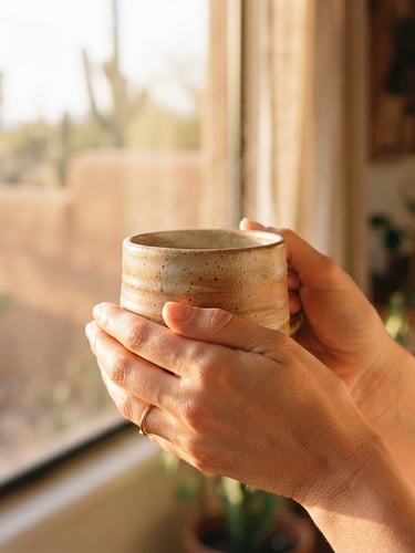 A close-up of hands holding a warm ceramic mug in soft golden-hour window light, muted desert earth tones, shallow depth of field, 35mm